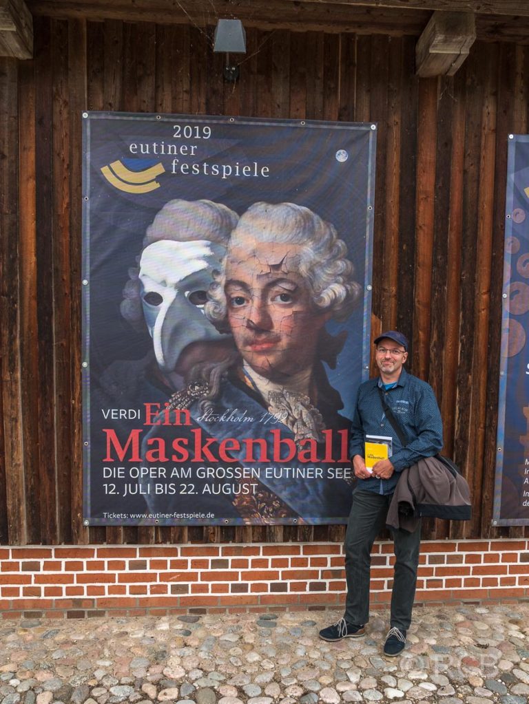 Mann vor Plakat der Eutiner Festspiele mit Verdis "Maskenball"