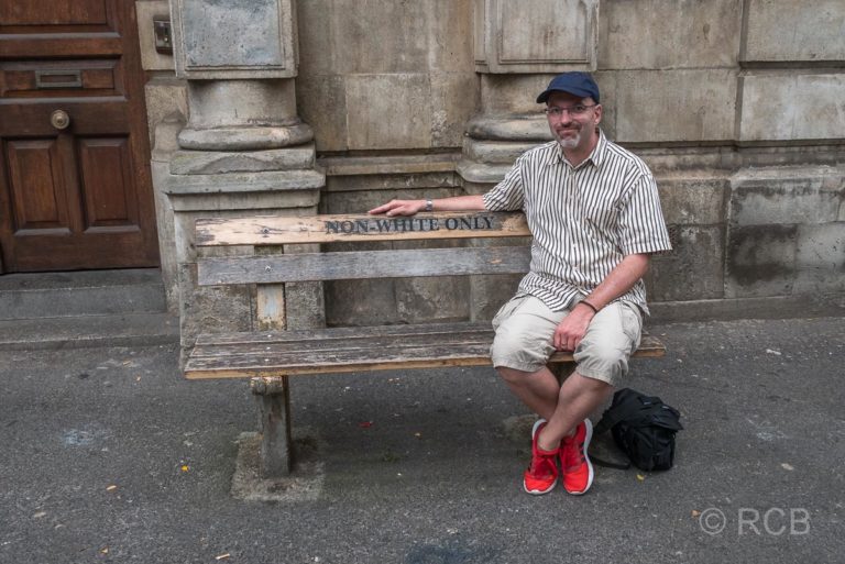 weißer Mann sitzt auf einer Bank aus der Zeit aus der Apartheid mit der Aufschrift "Nur für Nicht-Weiße"