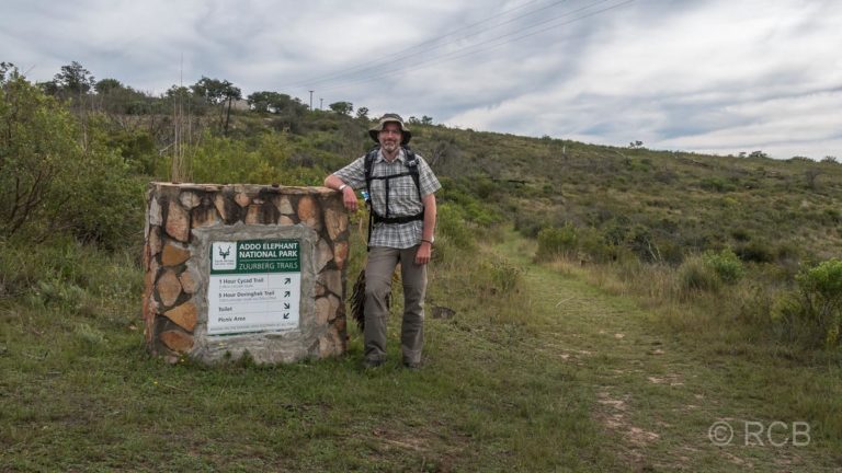 Mann lehnt an einem Schild, das den Beginn eines Wanderwegs im Addo Elephant National Park markiert
