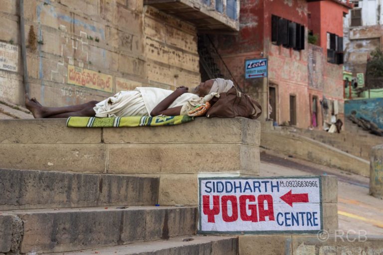 Varanasi, an den Ghats, Mann liegt entspannt auf den Stufen neben einem Werbeschild für ein Yoga-Zentrum