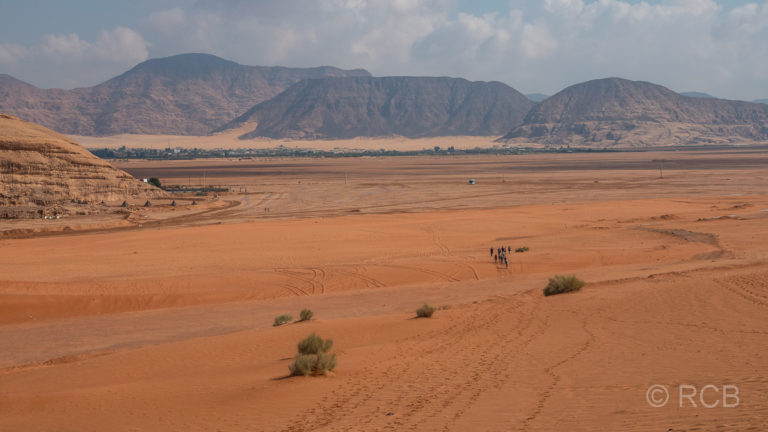 Wanderer laufen in der Ferne auf eine Siedlung am Rande des Wadi Rum zu.