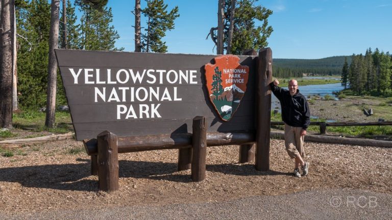 Mann lehnt an einem Schild am Südeingang des Yellowstone National Parks